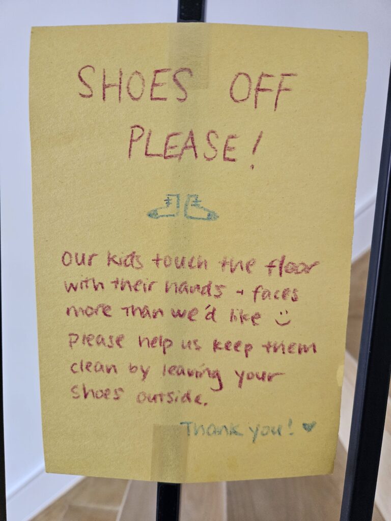 Handwritten note reads: Shoes off please! Our kids touch the floor with their hands + faces more than we'd like :) Please help us keep them clean by leaving your shoes outside. Thank you!